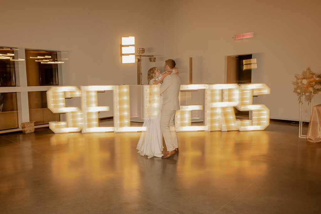 bride and groom first dance in front of light bulb name sign