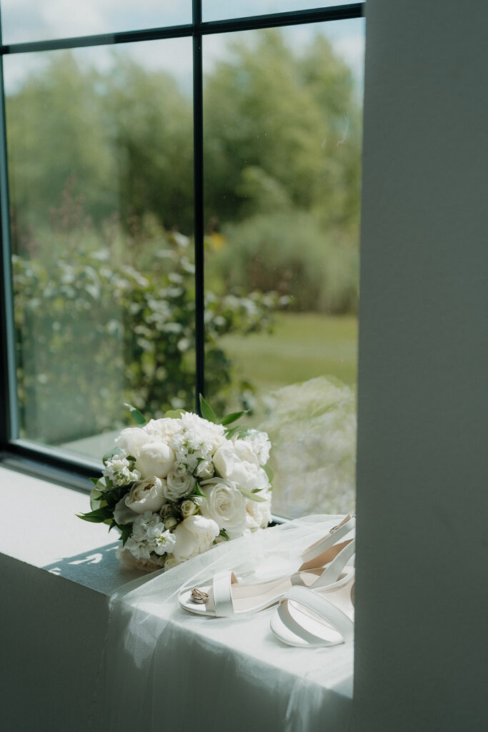 A flatlay photo of the brides shoes, veil, and bouquet resting on a window sill