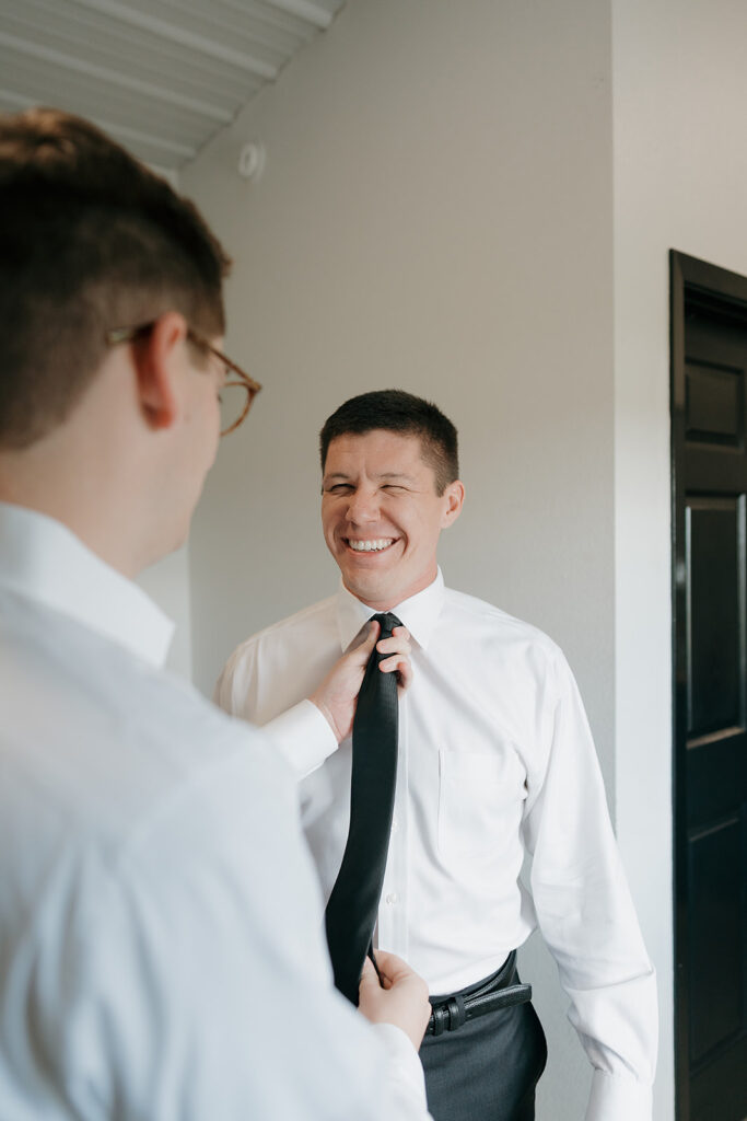 The best man helping the groom with his tie as they laugh together.