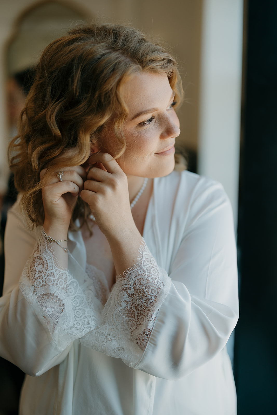 Bride adjusting her earrings near a window while getting ready, soft light highlighting her face — a quiet detail captured through documentary wedding photography
