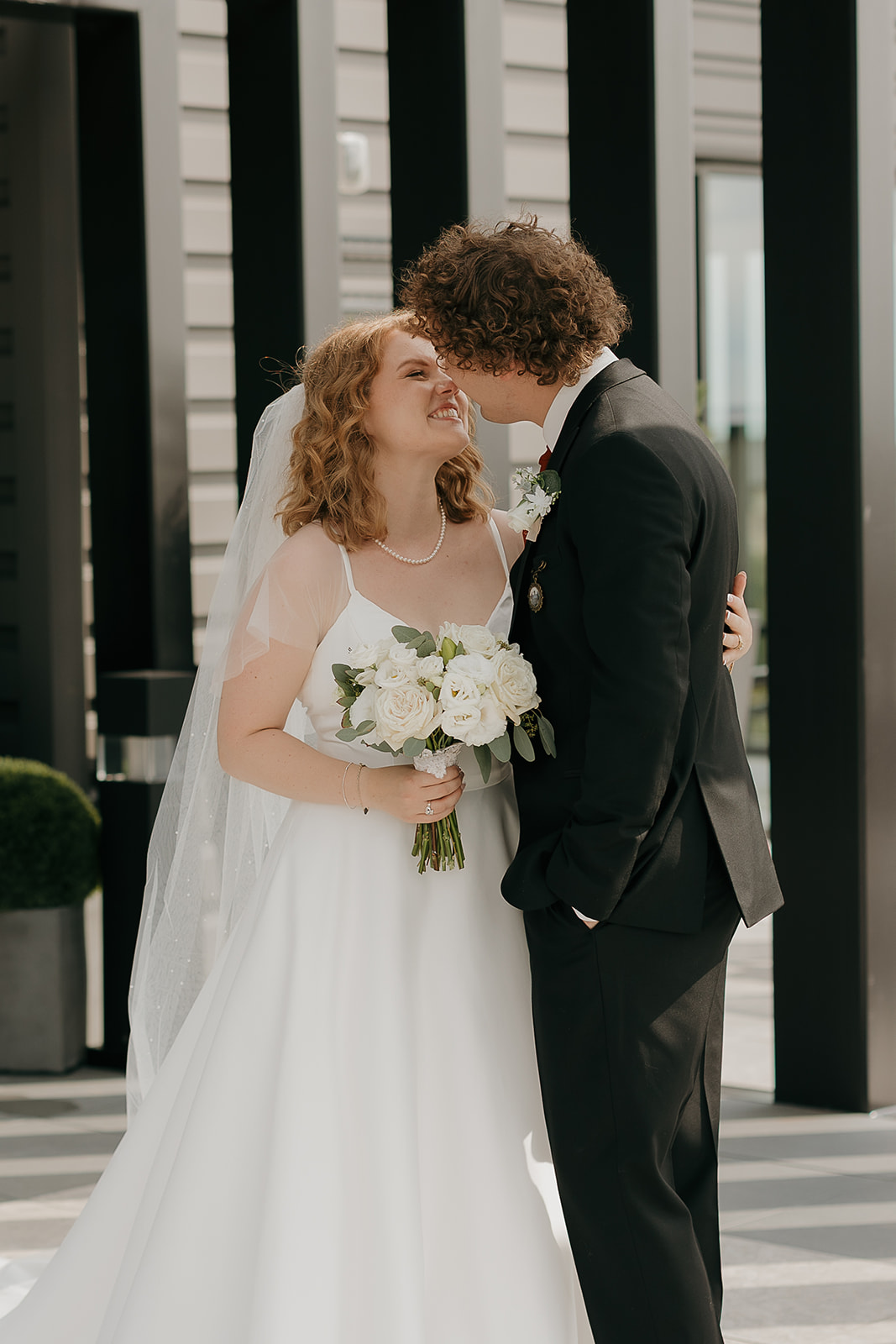 Bride and groom smiling and leaning in for a kiss while holding a white rose bouquet