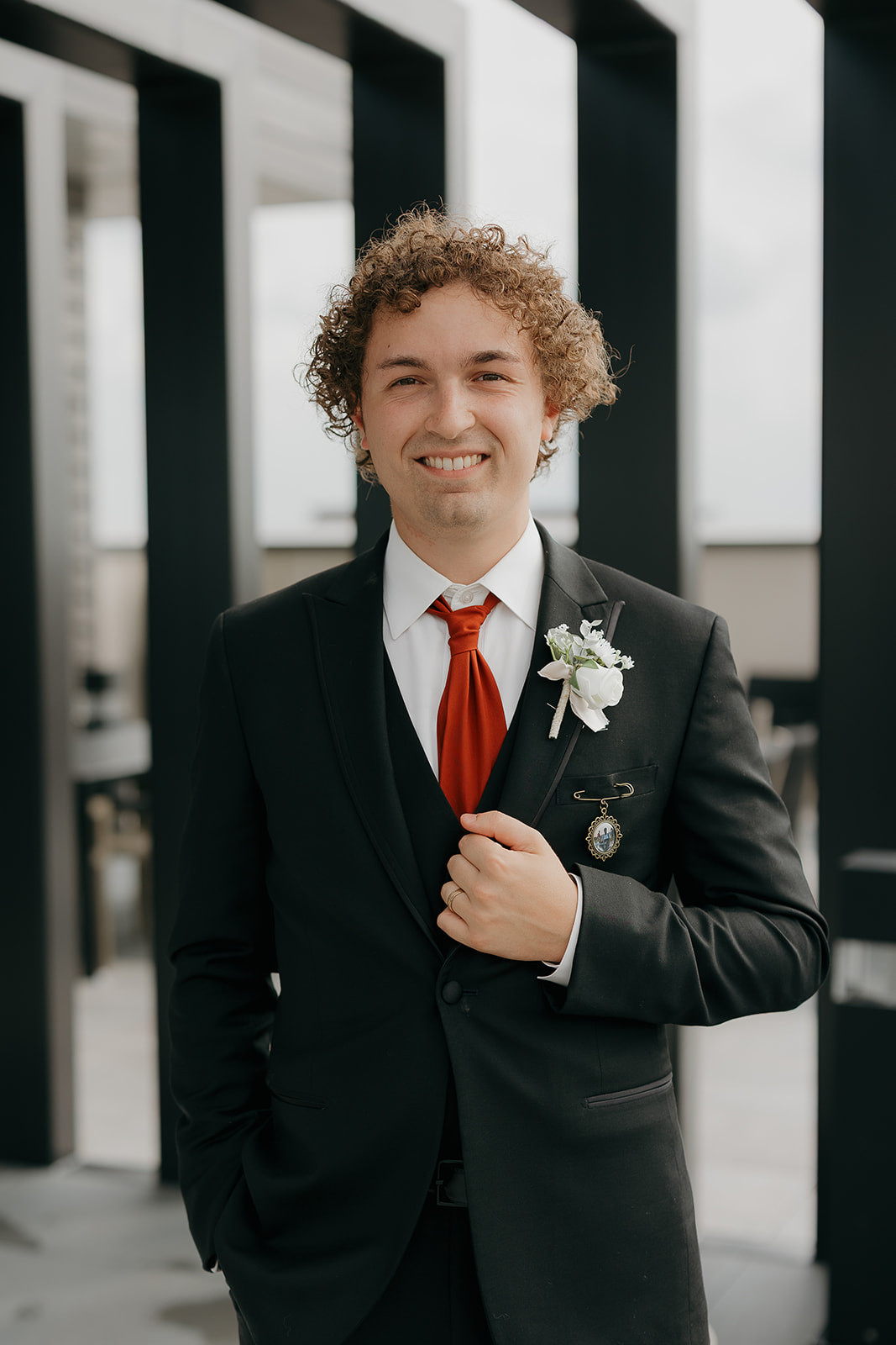 Groom standing outdoors in a black suit and red tie, smiling at the camera with his boutonniere