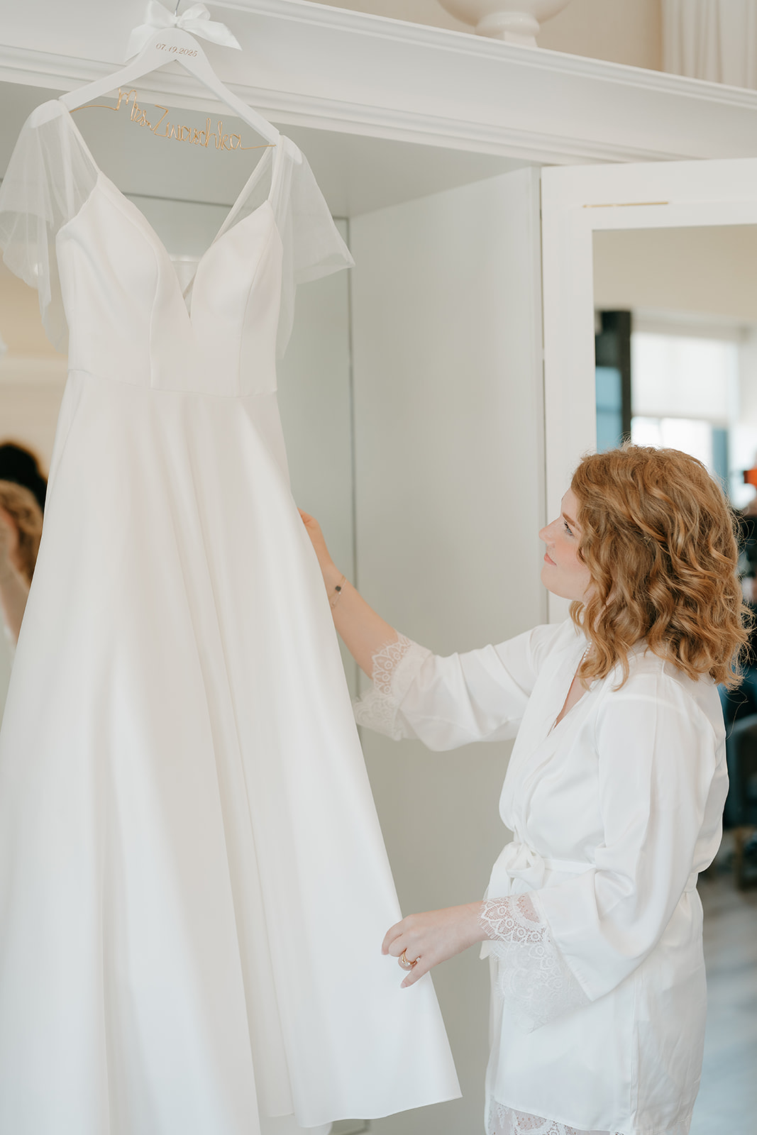 Bride touching her hanging wedding dress while wearing a white robe, getting ready before the ceremony