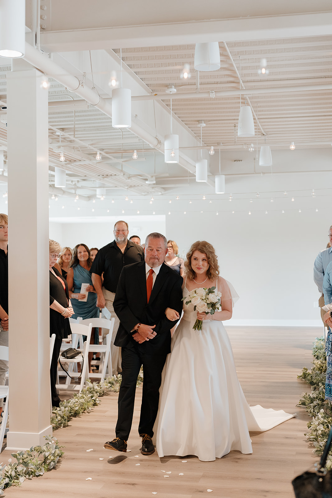 Bride walking down the aisle with her father, surrounded by guests and soft ceremony lighting — a meaningful moment captured through documentary wedding photography