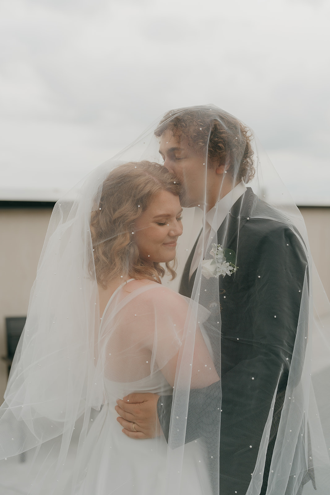 Bride and groom standing closely under the veil as he kisses her forehead outside