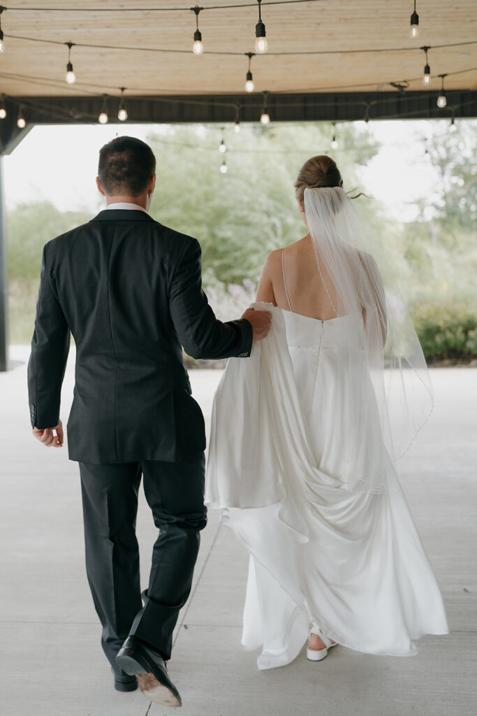 The groom is holding the brides train as they walk around their outdoor wedding venue in Iowa. 