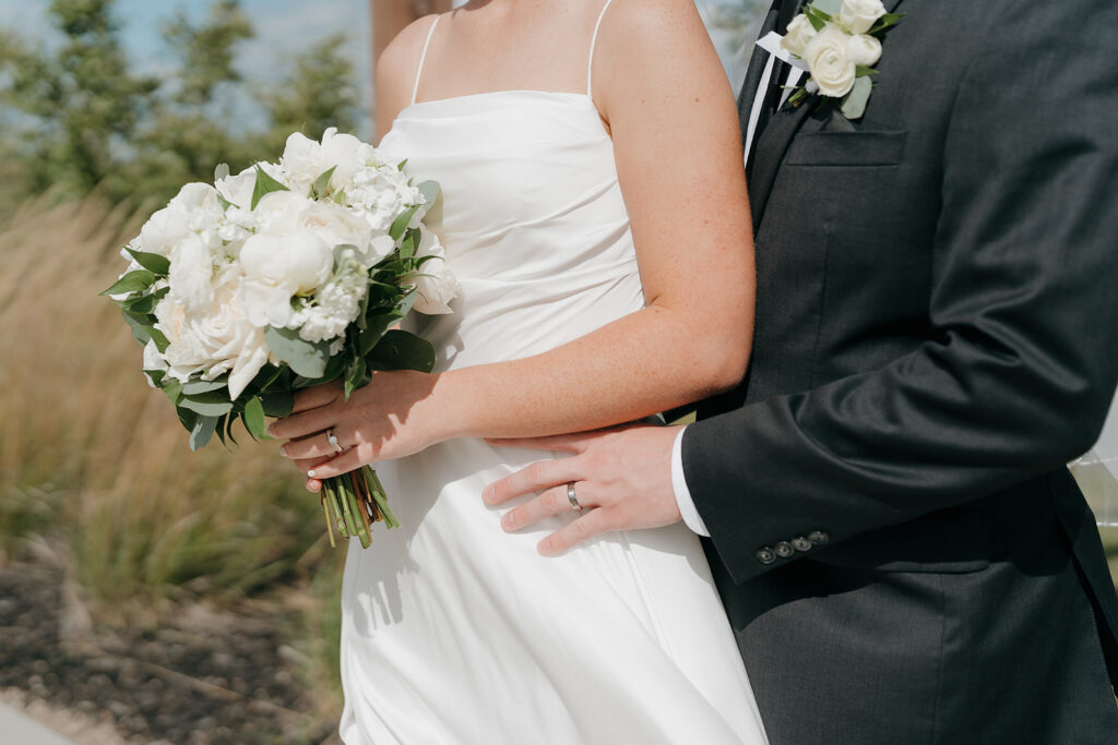 A horizontal detail shot of the bride holding her white bridal bouquet and the groom hugging her around the waist.