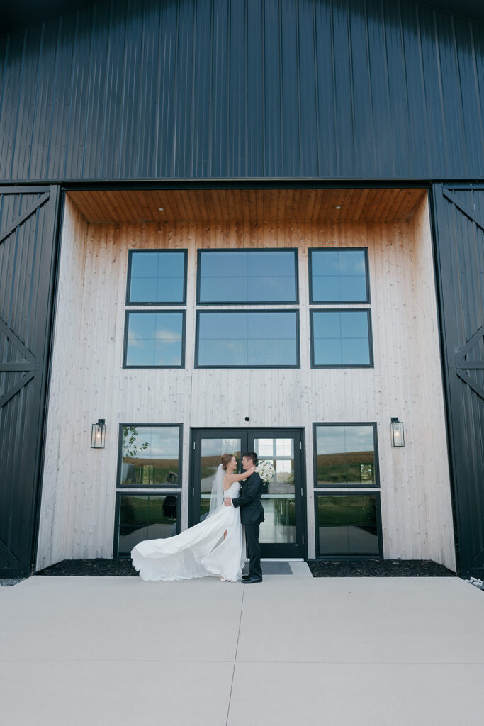 Wide photo of the bride and groom standing in front of the barn doors at their outdoor wedding venue for some bride and groom portraits