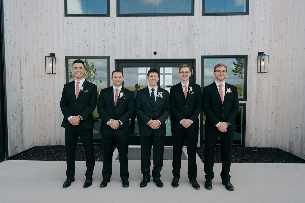 The groom and groomsmen lined up for groomsmen photos in front of their outdoor wedding venue in Iowa