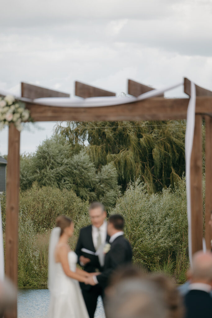 A cinematic shot of the bride and groom blurred out as they stand at the alter with the focus on the willow trees behind them through their wedding arch.
