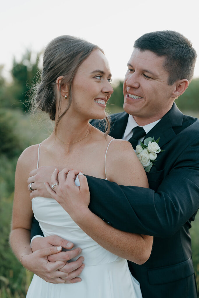 A stunning bride and groom portrait of the groom hugging around the bride from behind as they smile at each other during sunset.