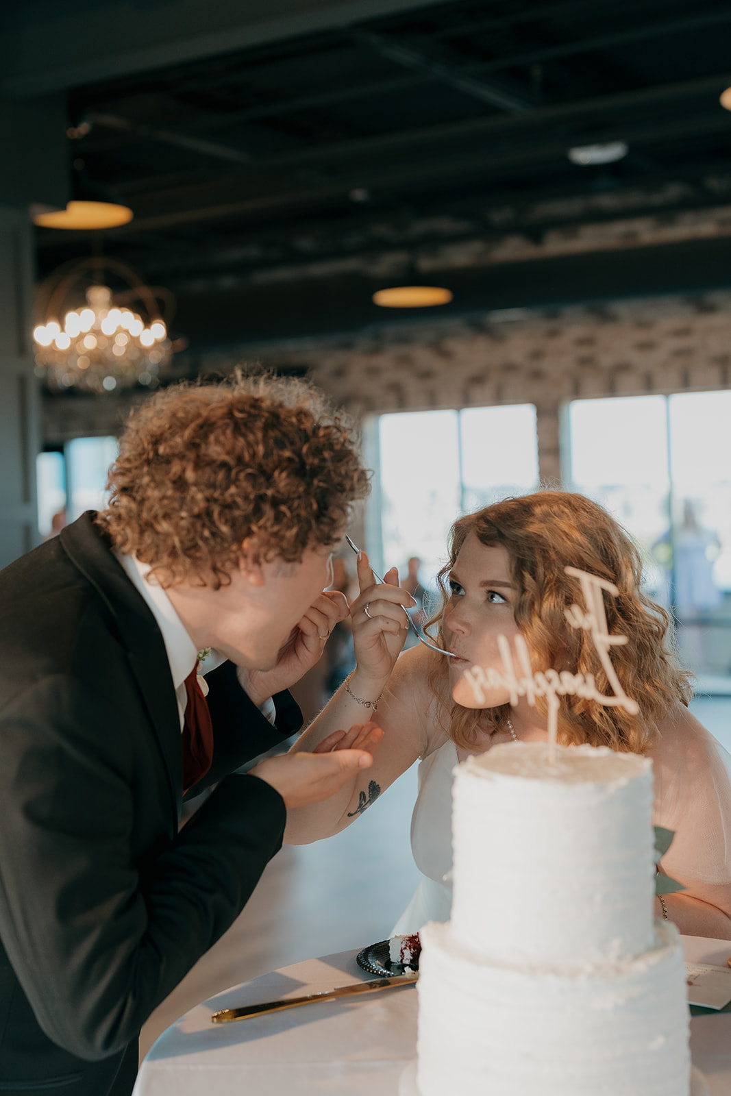 Bride and groom feeding each other cake during their reception, laughing together at the table