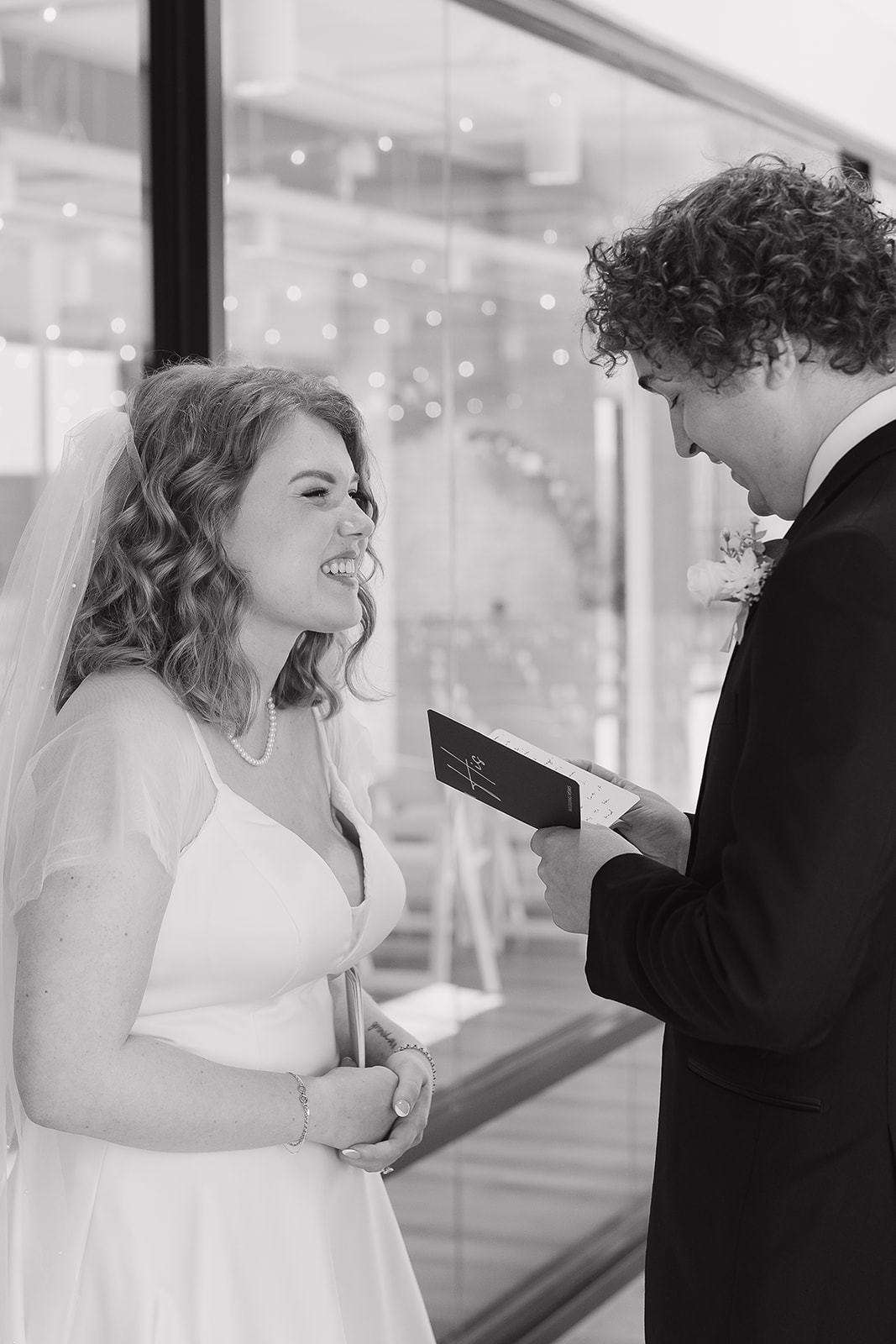 A black and white photo of the bride laughing at the groom while he reads his vows to her.