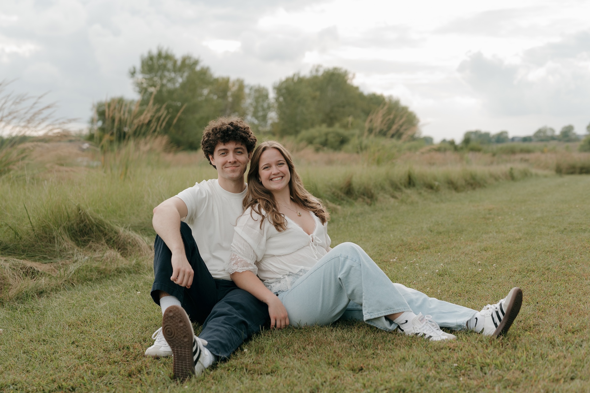 A wide shot of a couple sitting together on the grass in an open field, smiling at the camera during their engagement session.