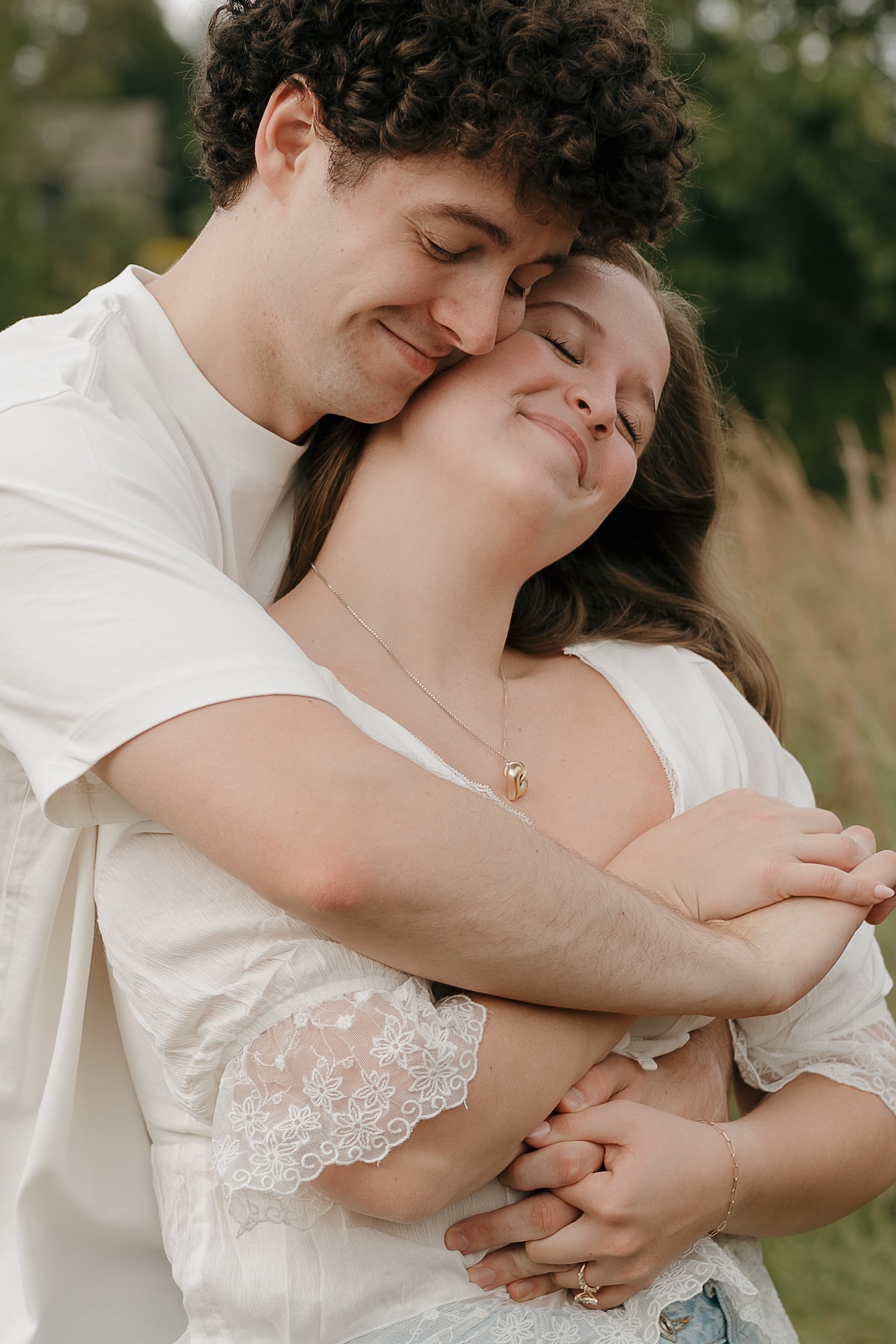 A couple hugging in a grassy field with tall trees behind them, highlighting peaceful outdoor Engagement Photo Locations.
