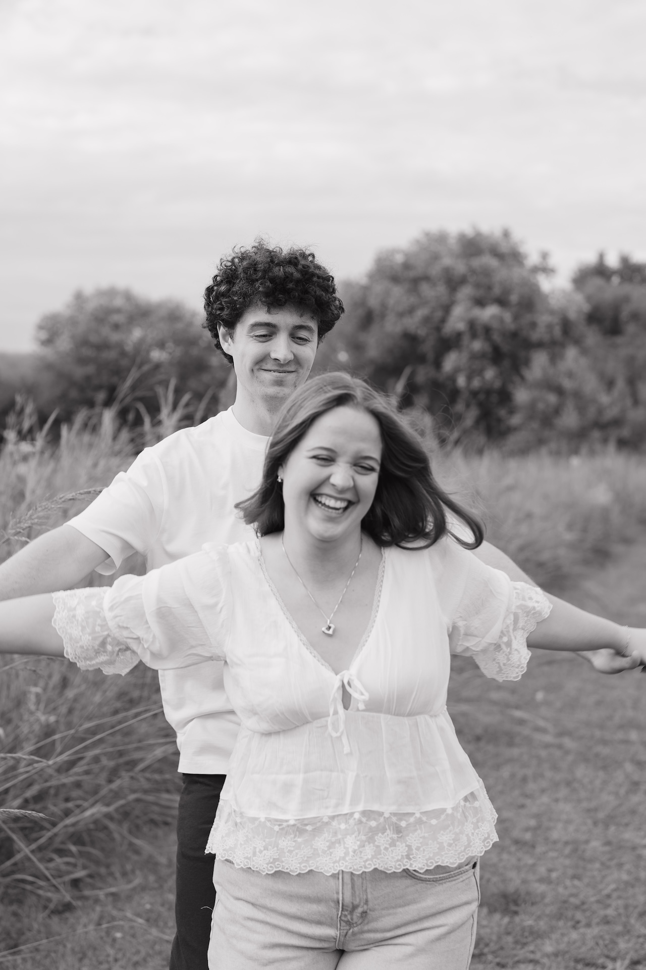 Black and white photo of a couple laughing together while walking through an open field, arms outstretched and enjoying the moment.