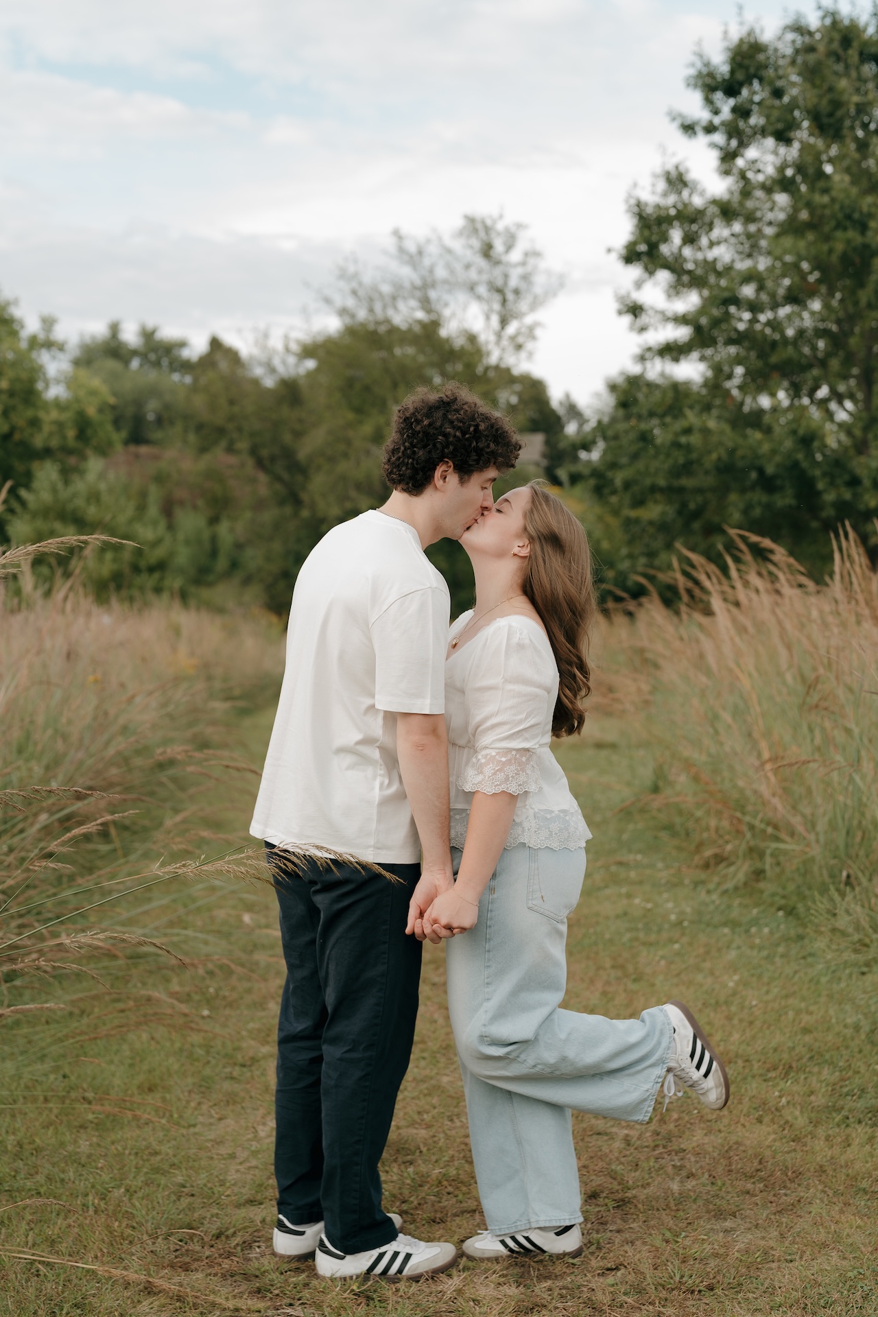 Wide shot of a couple kissing while holding hands in a grassy field, highlighting natural Engagement Photo Locations with tall grasses and open space.