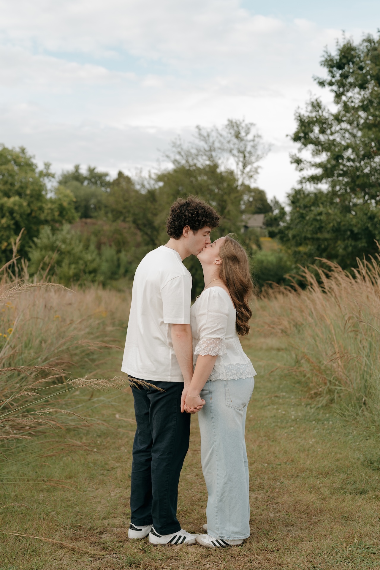 Couple sharing a kiss while holding hands on a narrow path surrounded by tall grass, captured during a natural engagement photo session outdoors.