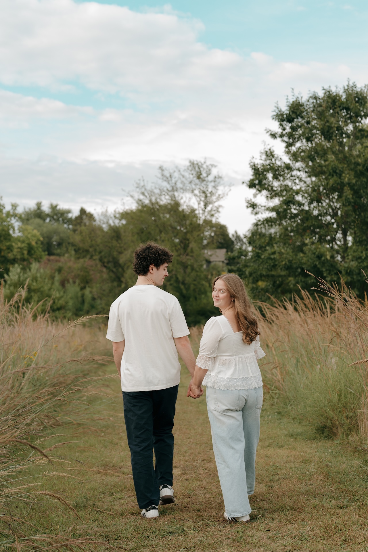 Engagement photo of a couple walking hand in hand away from the camera through tall grass, with trees and open sky in the background.