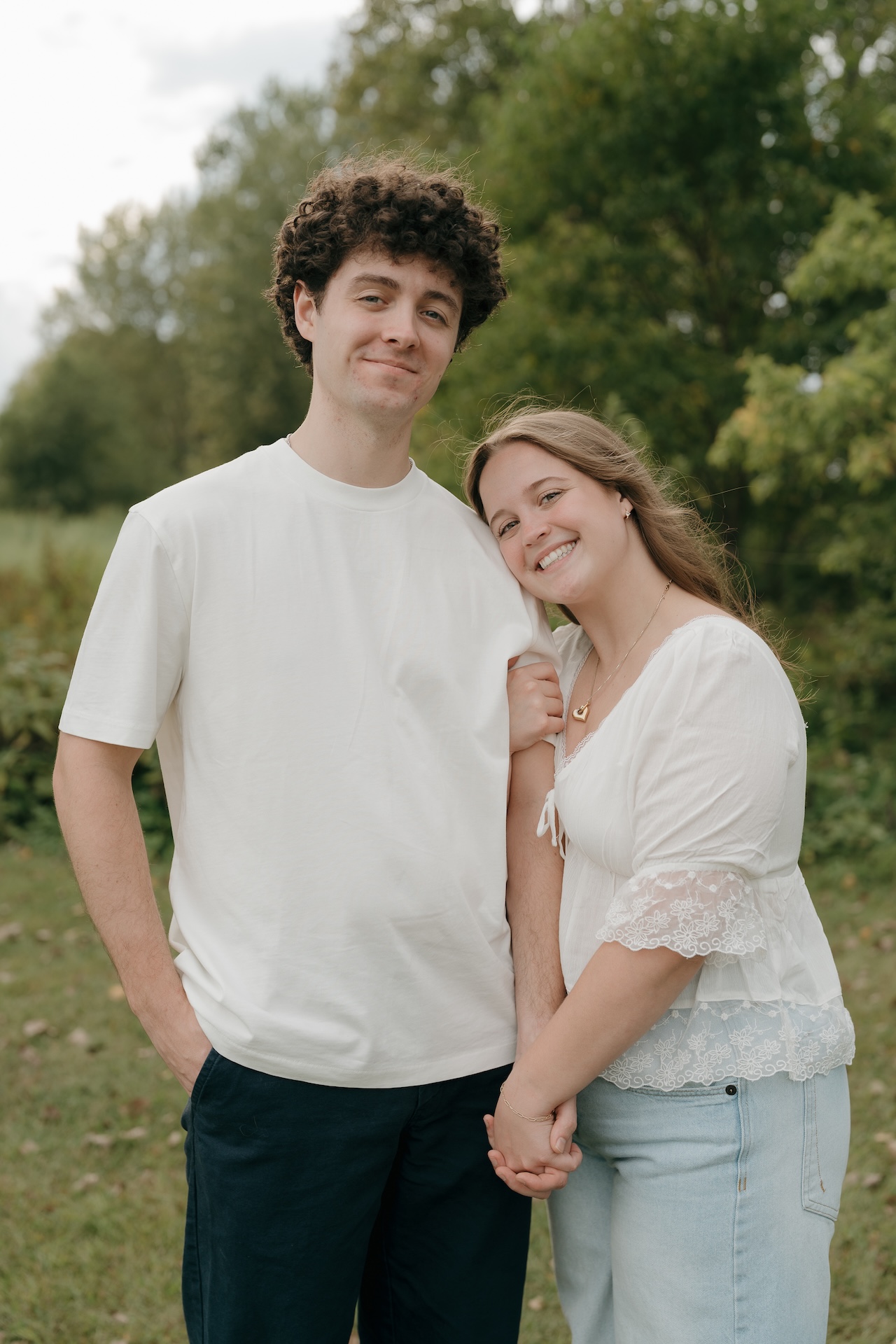 Couple standing side by side in a grassy field, smiling at the camera while holding hands during a relaxed outdoor engagement session.