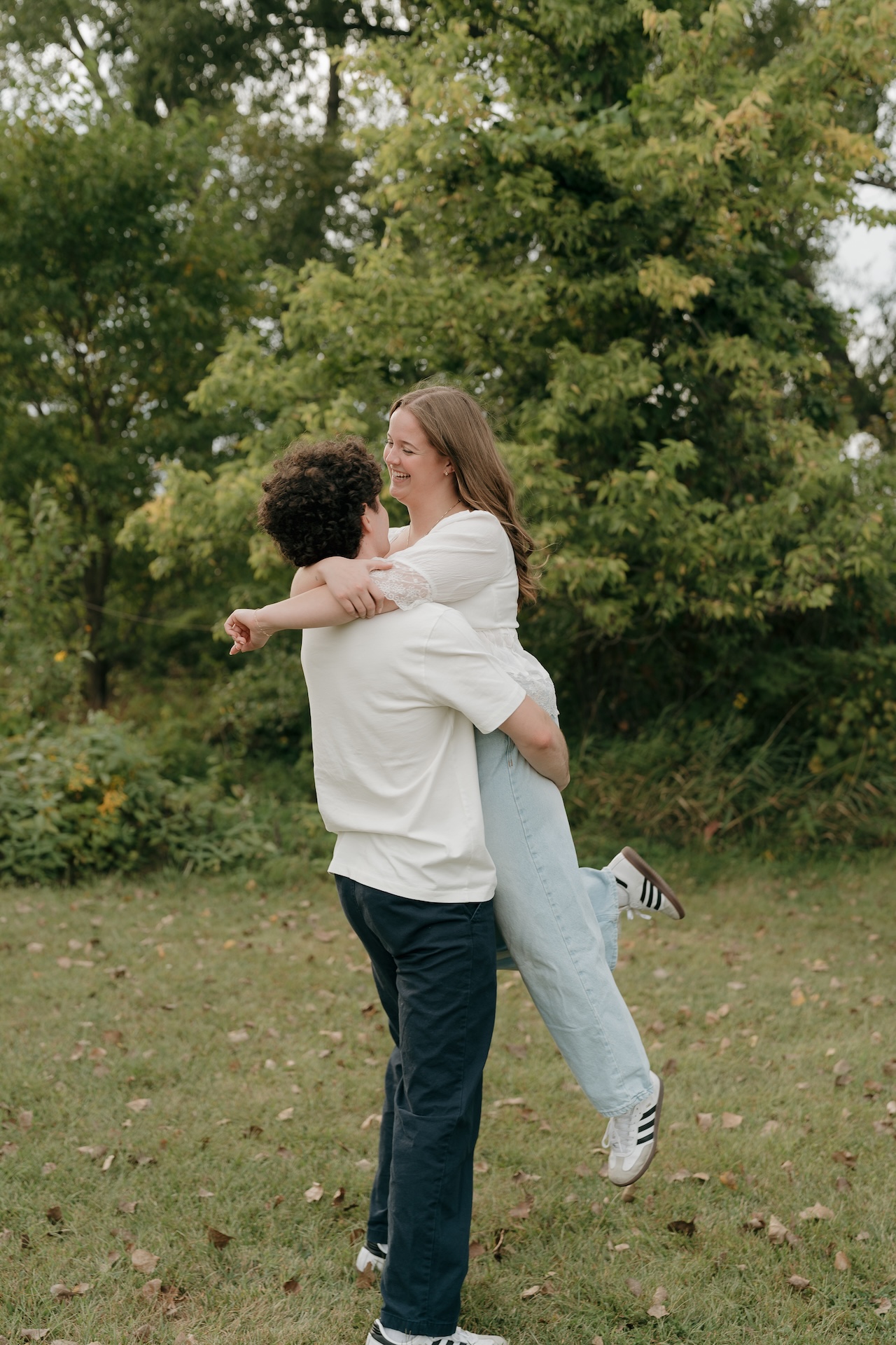 A couple hugging and laughing in a grassy field surrounded by trees, captured at one of their favorite Engagement Photo Locations.