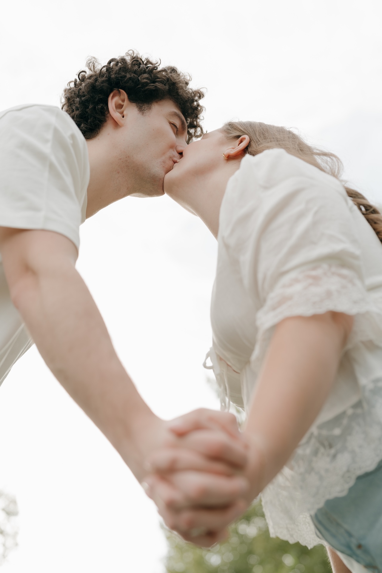Low-angle photo of a couple kissing while holding hands, with open sky above them creating a soft and romantic feel.
