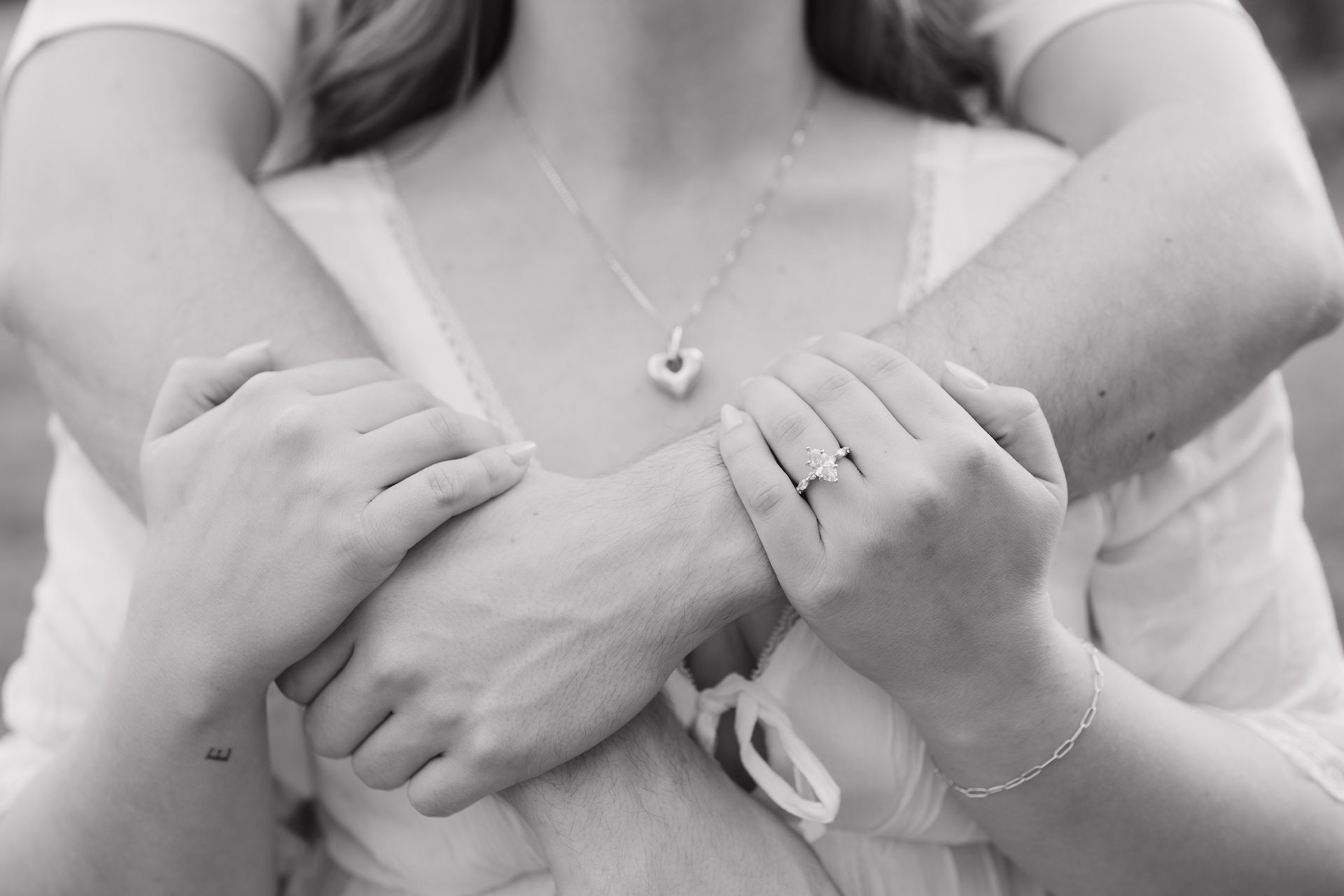 Black and white close-up of a couple embracing from behind, focusing on their arms, hands, and subtle jewelry details.