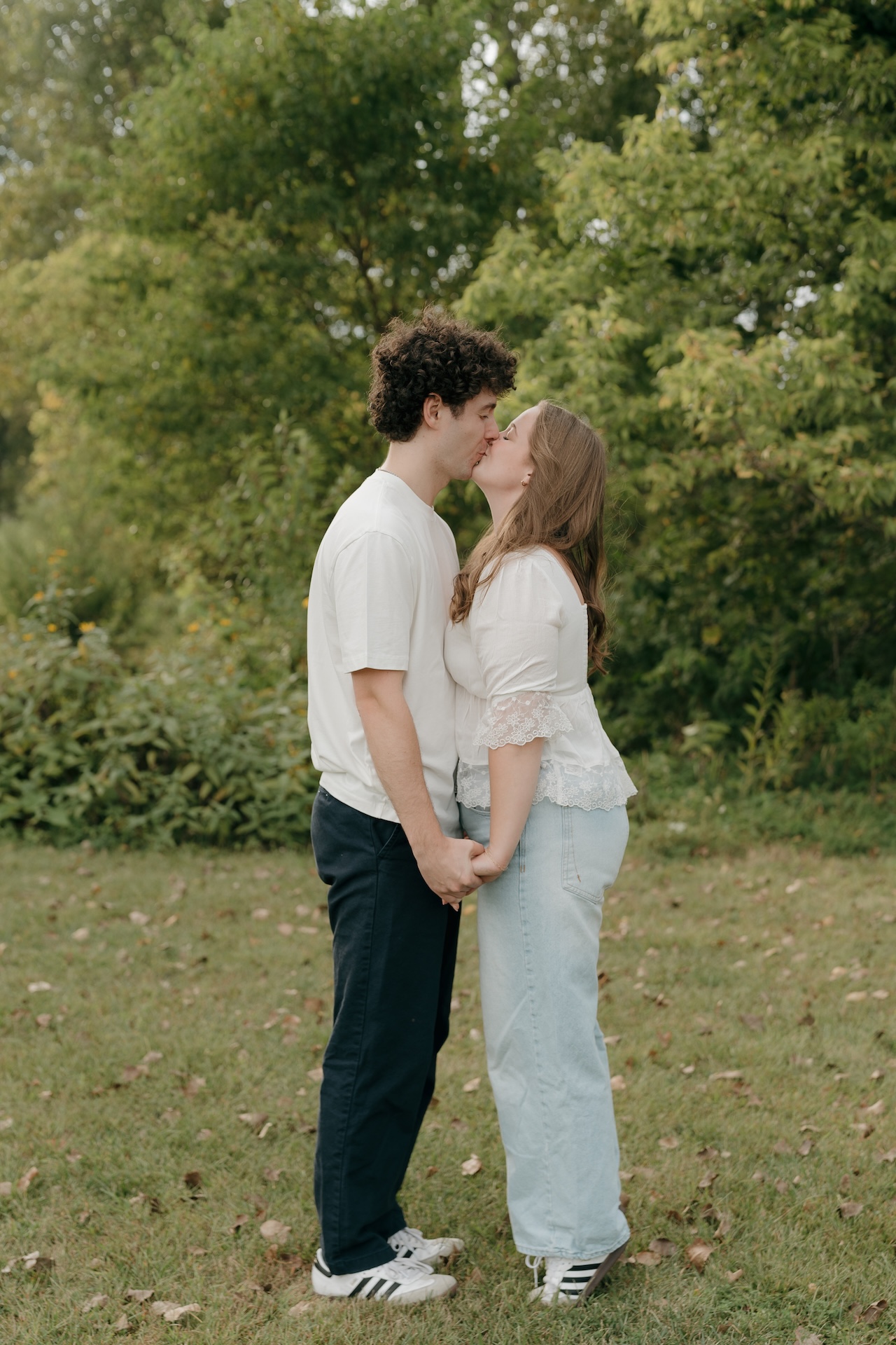 A couple standing face to face and kissing while holding hands, photographed at scenic Engagement Photo Locations surrounded by greenery.