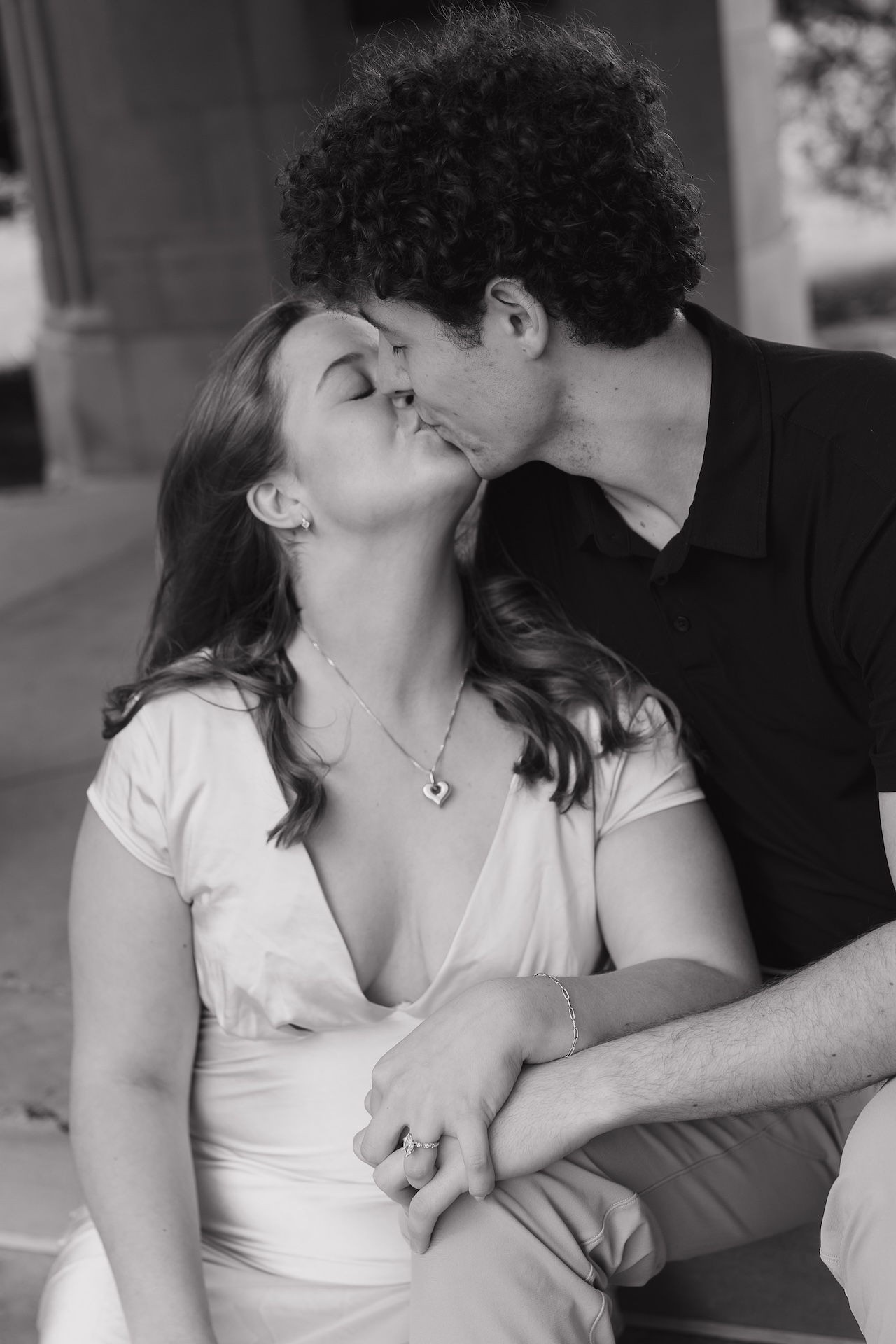 Black and white photo of a couple kissing while holding hands in front of a large stone building with tall arched windows.
