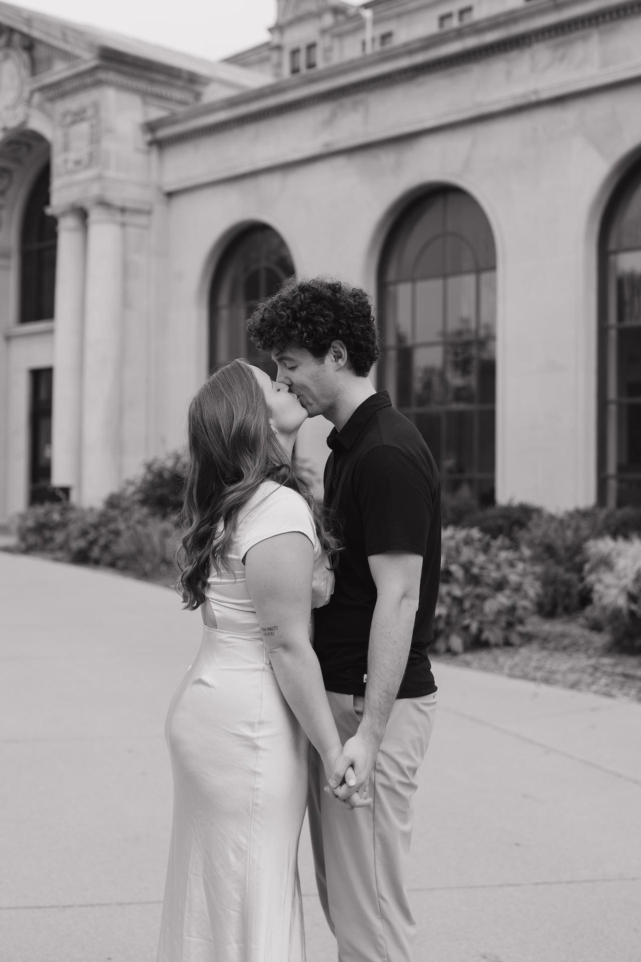 Black and white photo of a couple kissing while holding hands in front of a large stone building with tall arched windows.