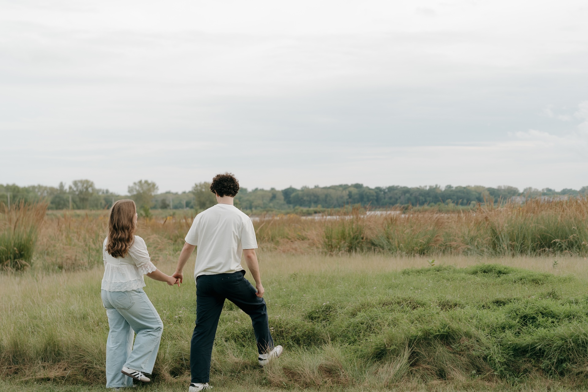 Engagement photo of a couple walking hand in hand across a grassy landscape, stepping over uneven ground with a wide open field behind them.