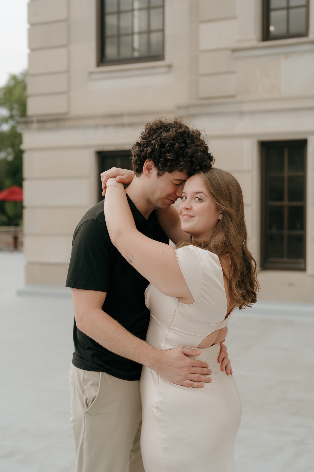 A couple embracing outdoors near an elegant building, with the woman looking over her shoulder and smiling during a relaxed engagement portrait.