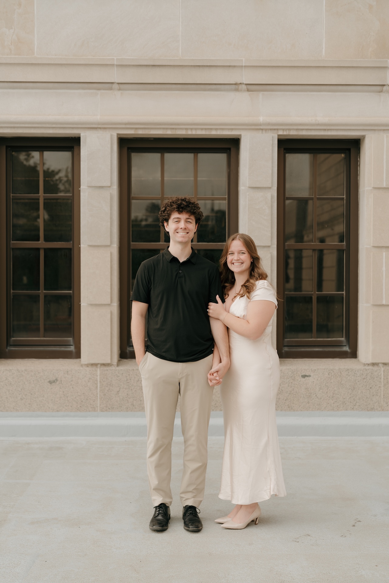 A couple standing hand in hand in front of a classic stone building with tall windows, smiling directly at the camera during their engagement session.