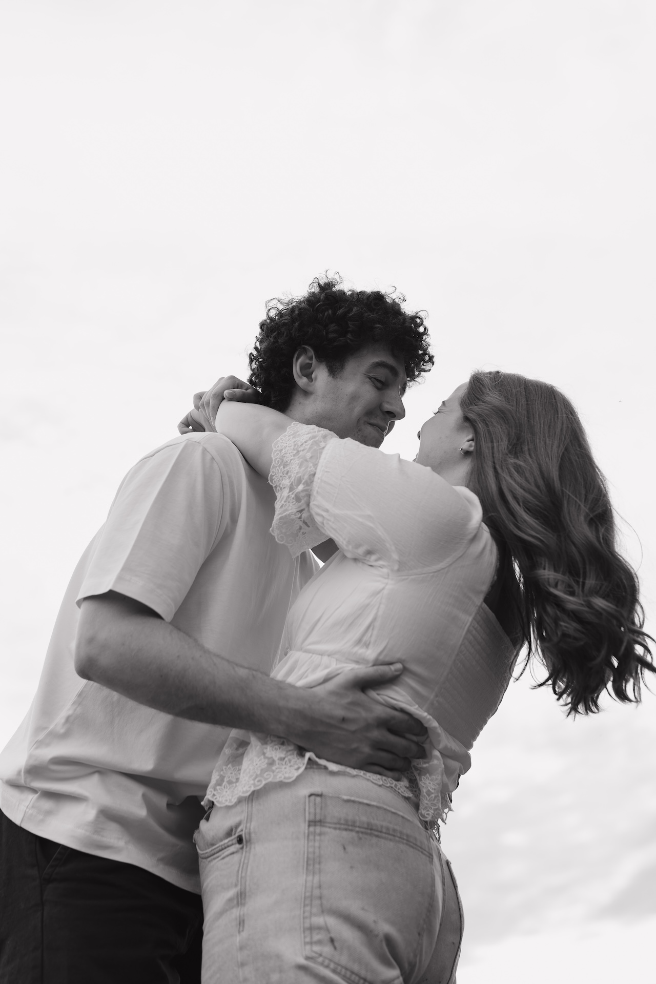 Low-angle black and white photo of a couple hugging and smiling at each other against an open sky during an engagement session.