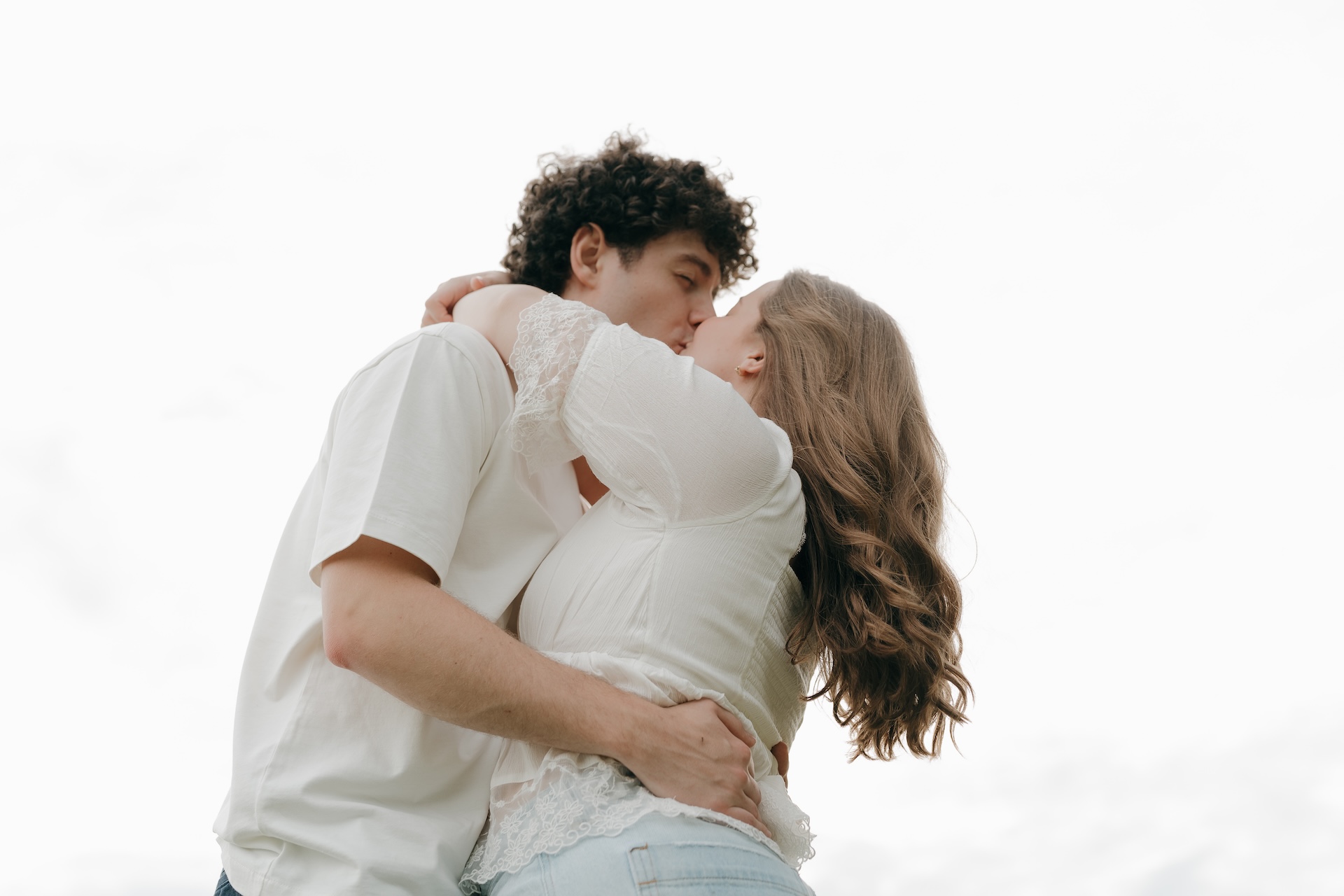 Low-angle engagement photo of a couple kissing against an open sky, with the bride wrapping her arms around the groom’s shoulders.