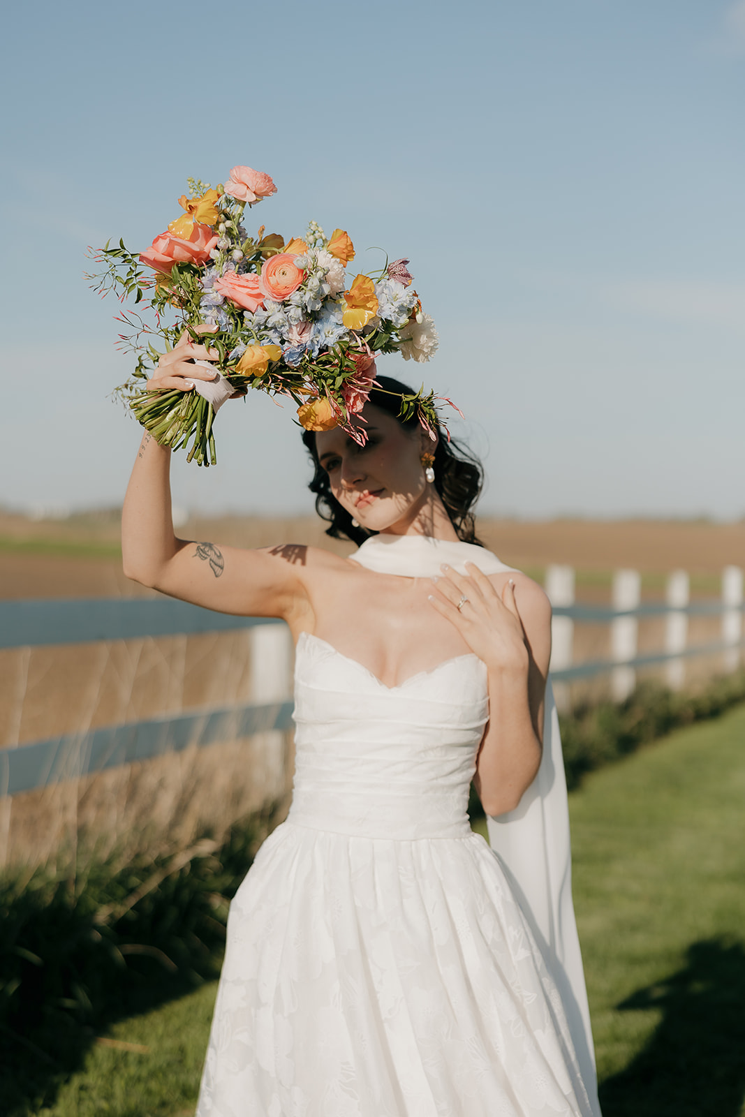 An editorial photo of a bride holding up her bright wedding bouquet to block the sun on her face while standing in a field.