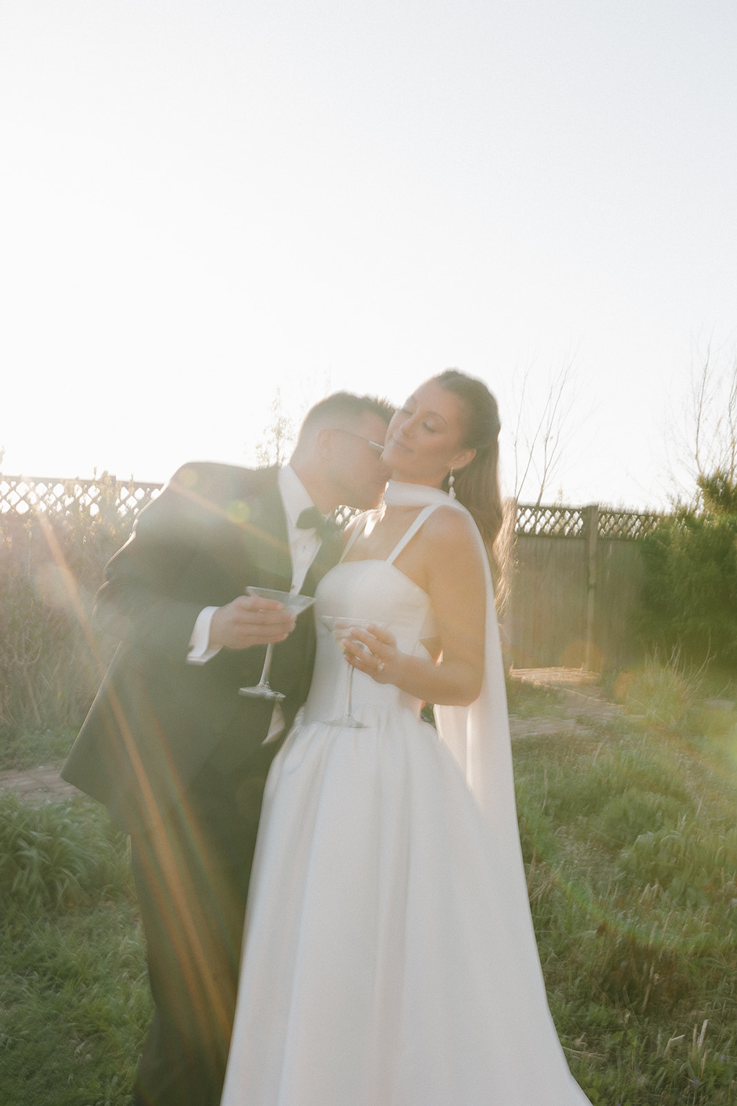Bride and groom standing in a grassy field during golden hour while holding martinis, with the groom kissing the bride’s cheek in soft backlit sunset light for a romantic outdoor wedding portrait.