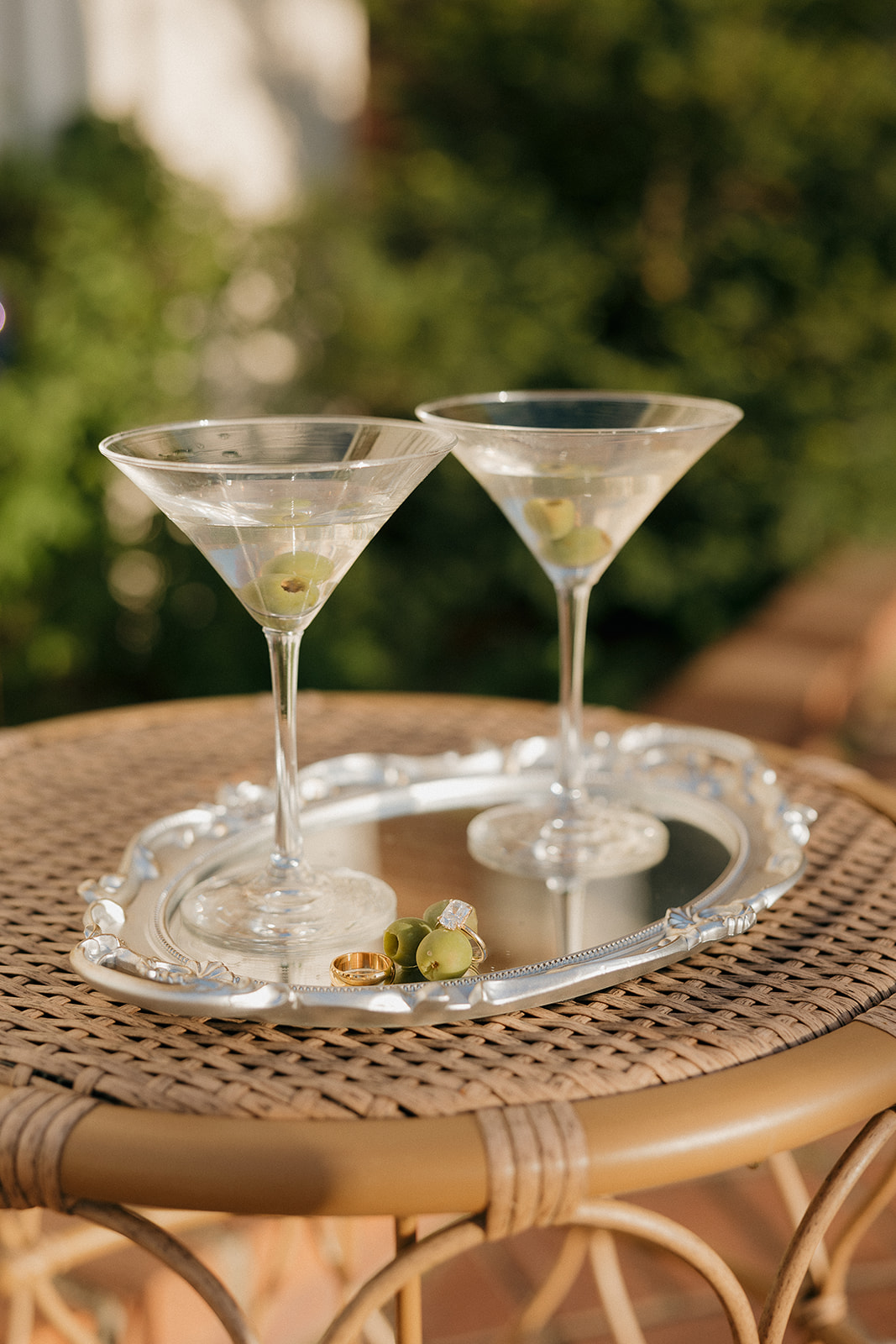 Two martini glasses with green olives on a silver tray beside wedding rings on a wicker table, styled using a wedding vendor checklist for cocktail hour details and flat lay inspiration.