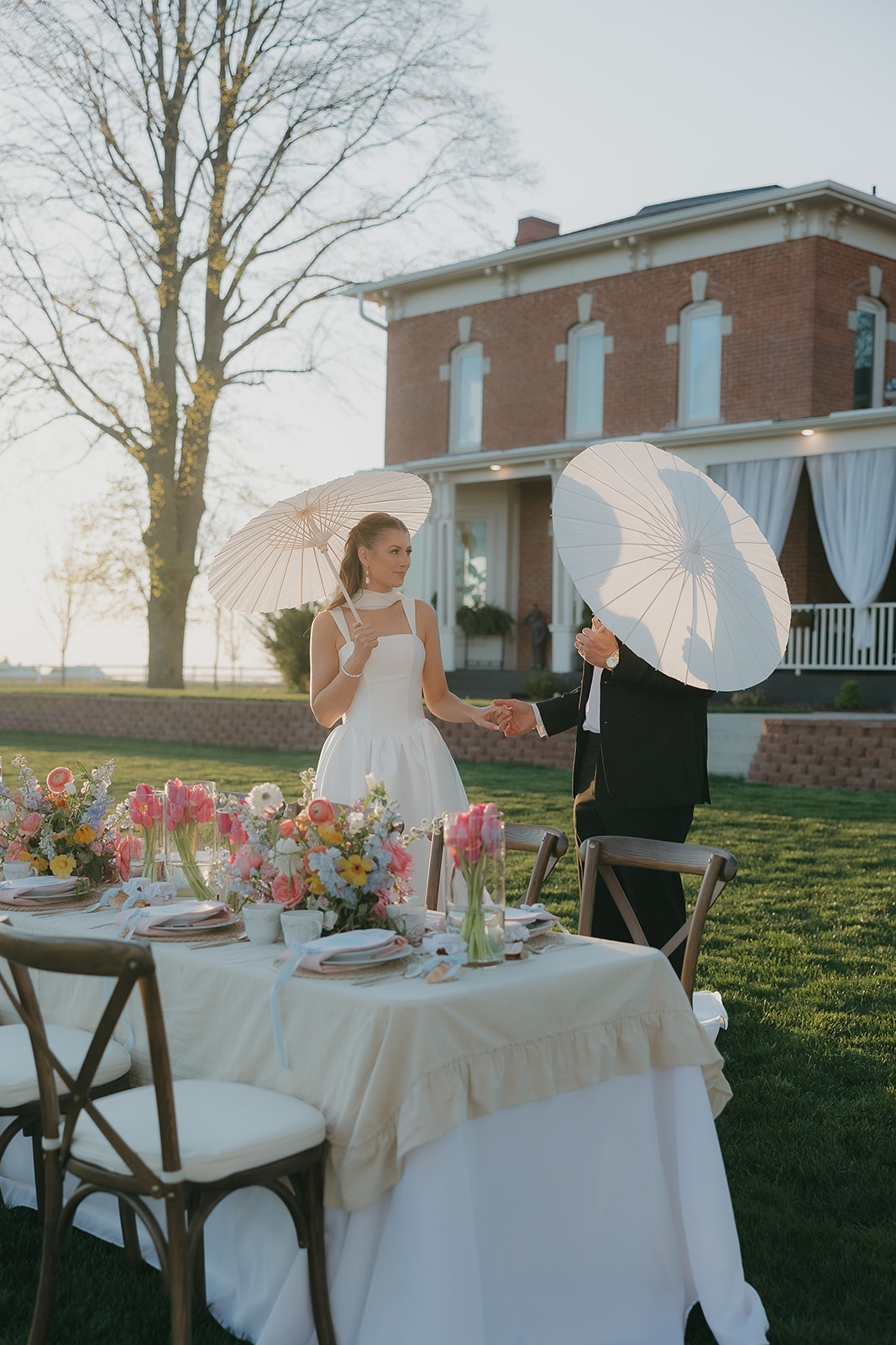 A bride and groom holding parisols during golden hour and holding hands in front of their wedding tables.