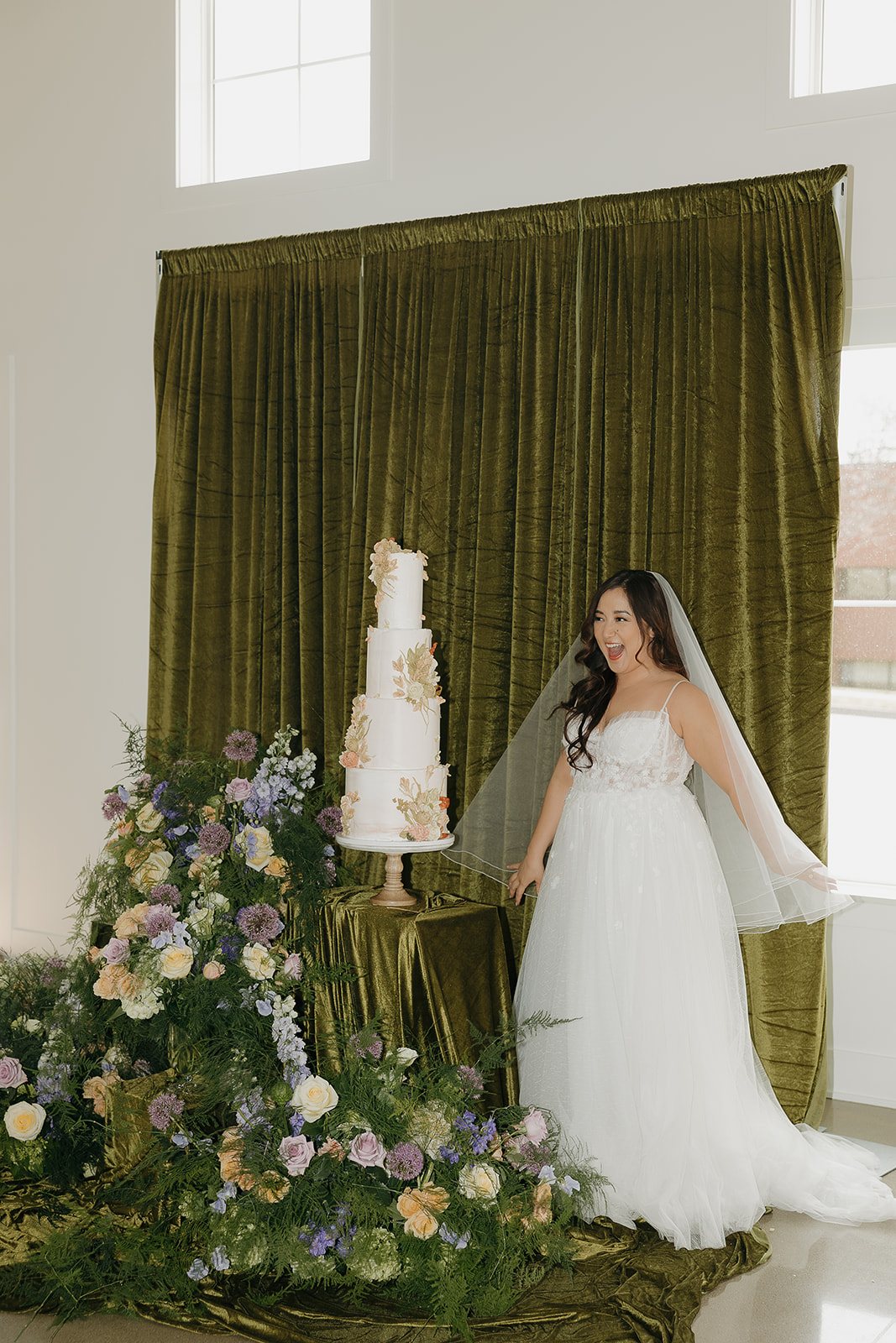 Bride standing beside a three-tier white wedding cake with floral appliqué in front of a green velvet backdrop, inspired by a wedding vendor checklist for cake and décor styling.