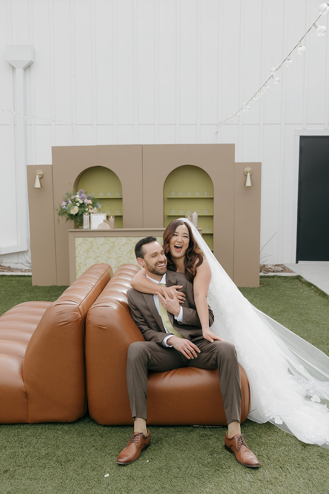 Bride sitting on a caramel leather couch while groom sits in front of her, both laughing during a playful editorial wedding portrait.