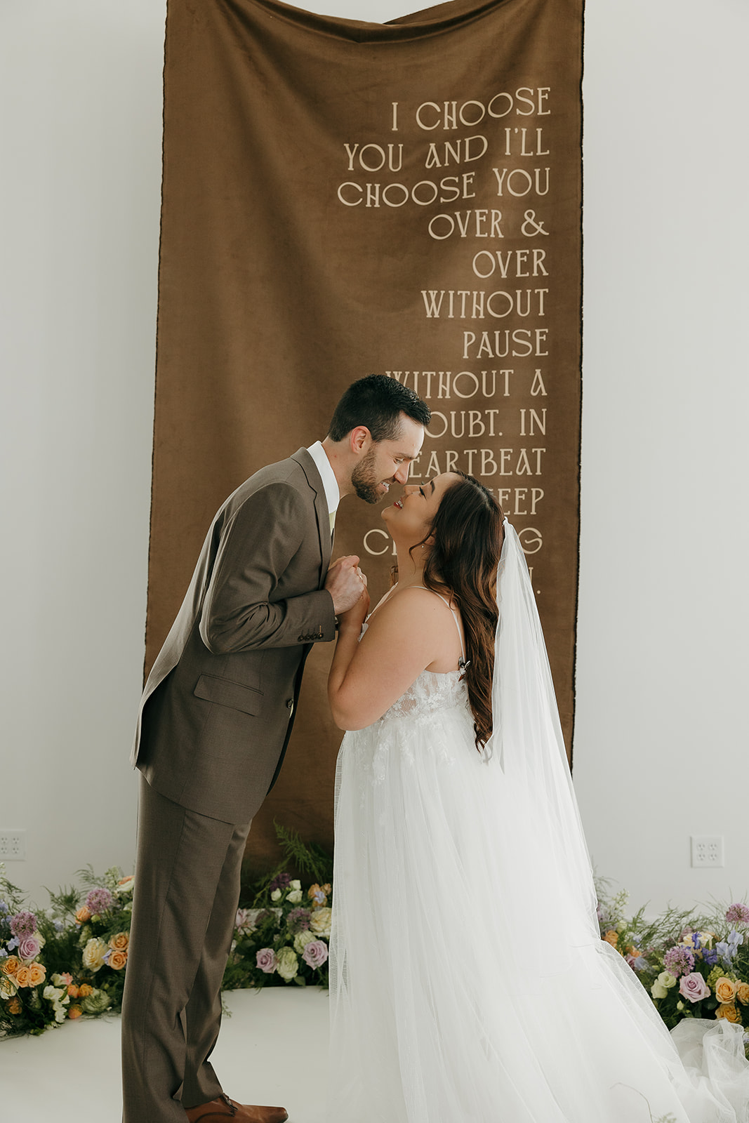 Bride and groom standing under a large brown fabric backdrop with romantic text during a styled indoor wedding portrait.
