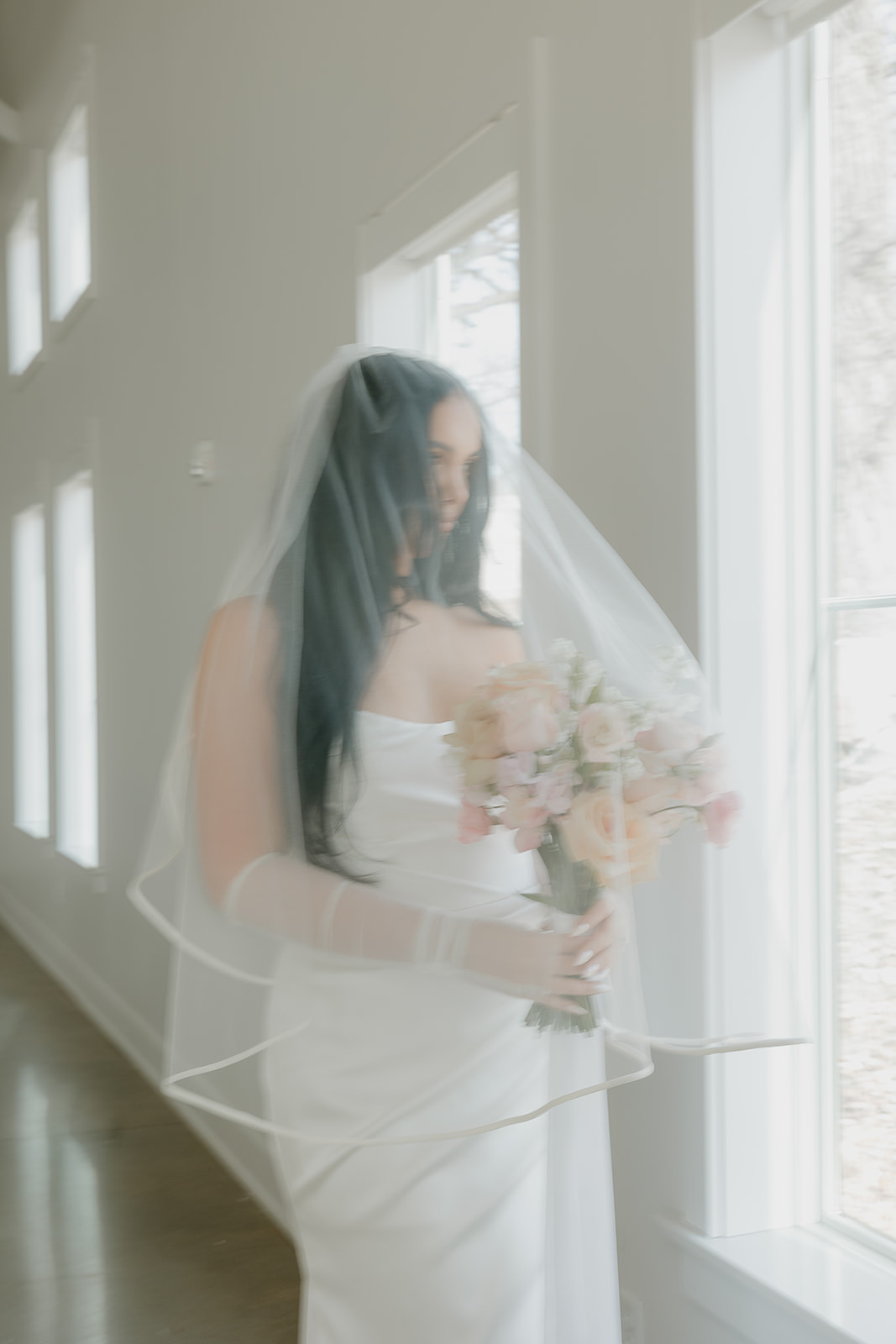 Bride walking through a bright white hallway in a strapless wedding dress and veil while holding a soft blush bouquet, inspired by a wedding vendor checklist for bridal portraits and timeline flow.