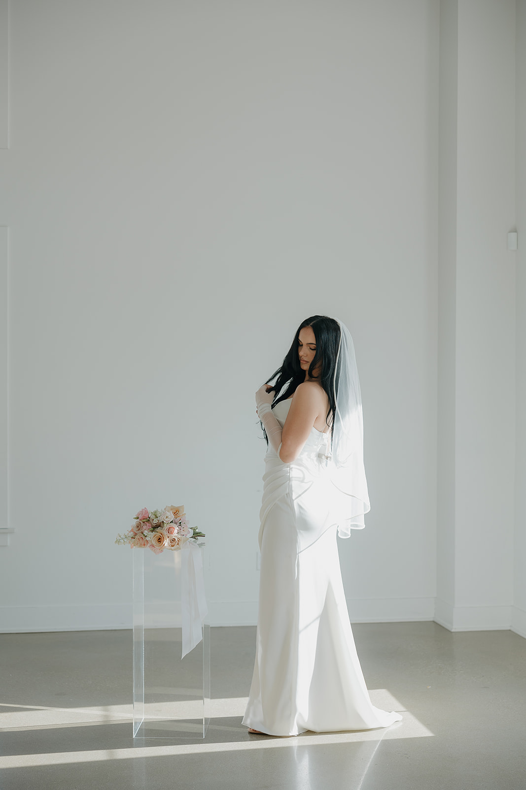 Minimalist bridal portrait of a bride standing in a sunlit white studio space with her bouquet resting on a clear pedestal, creating a modern editorial wedding aesthetic.