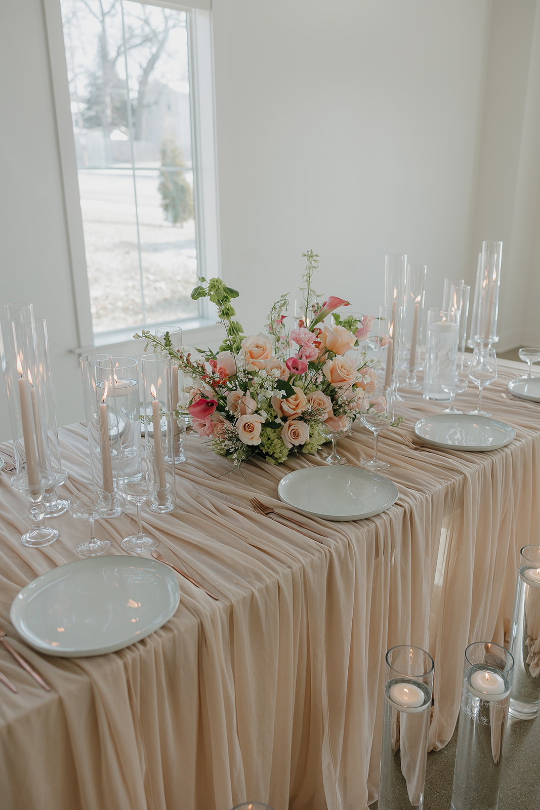 Long reception table with blush linens, pastel floral centerpiece, taper candles, and layered place settings in a bright white wedding venue interior.