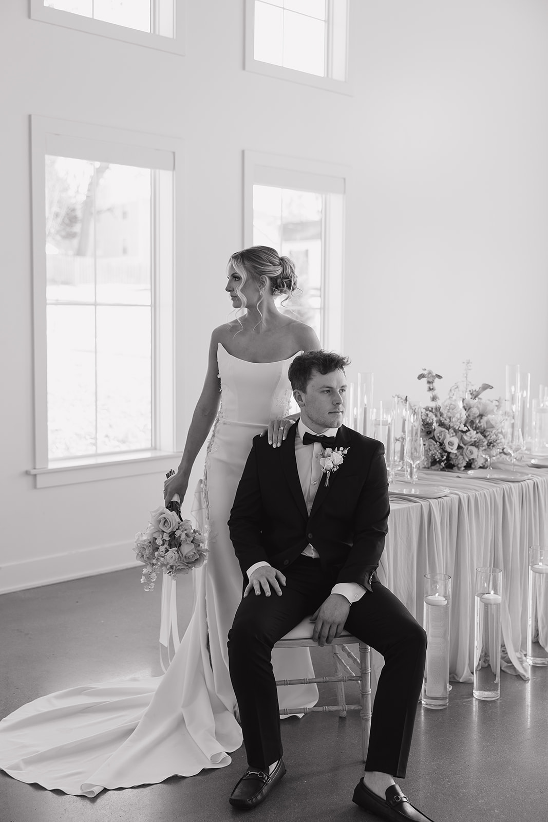 Black and white editorial wedding portrait of a bride standing behind the groom seated in a chair beside a candlelit reception table with florals and linens.