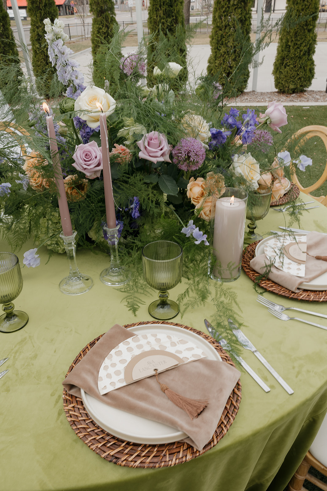 Pastel spring wedding tablescape with pink and lavender florals, green velvet linens, taper candles, and layered place settings for an elegant outdoor reception.