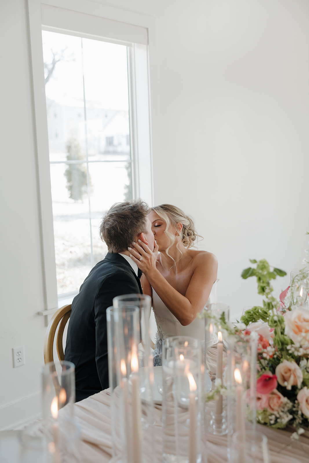 Bride kissing groom while seated at a candlelit reception table with pastel florals and layered place settings in a bright white venue.