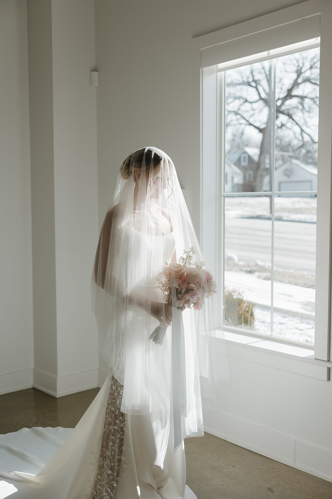 Bride standing by a window holding a bouquet under a long veil in natural light, inspired by a wedding vendor checklist for bridal portrait timing and lighting.