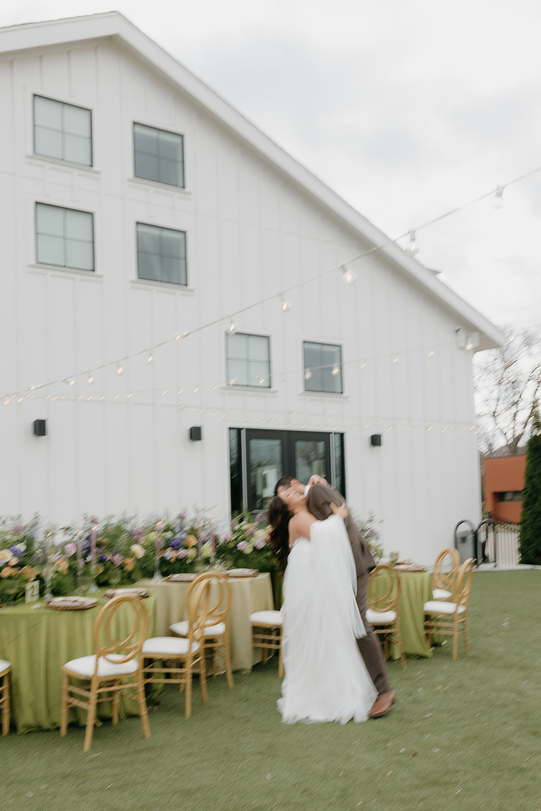 Bride and groom embracing in front of a white barn-style venue with long reception tables and string lights, inspired by a wedding vendor checklist for outdoor wedding layouts.